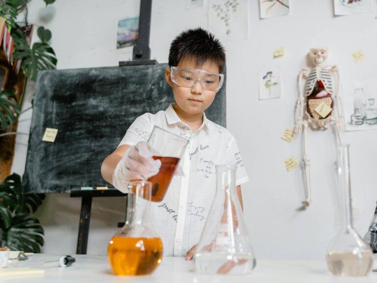 A child wearing goggles performs a chemistry experiment with colored liquids in a classroom.
